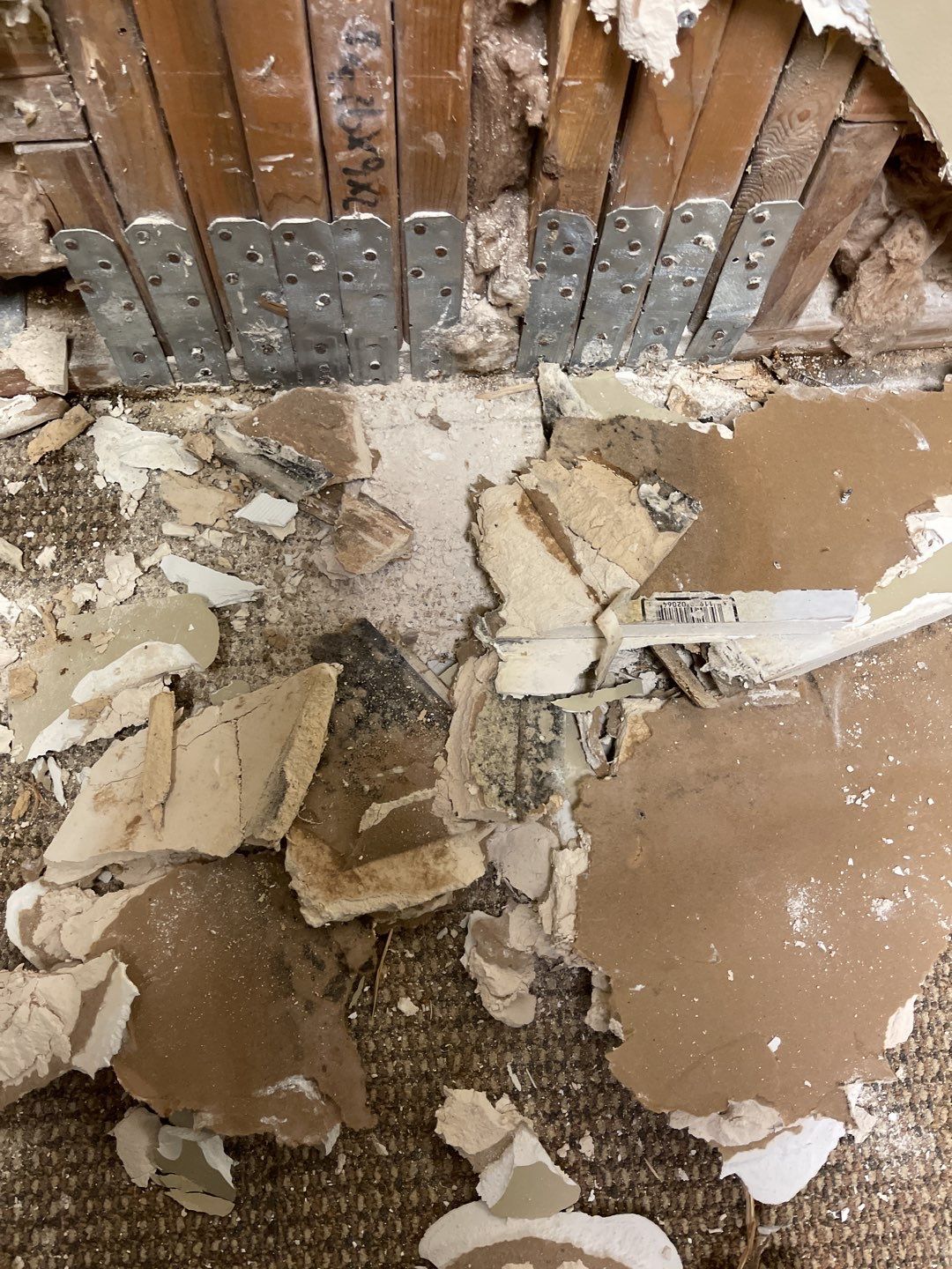 A close-up view shows a construction site with damaged drywall and exposed wooden studs. Metal brackets are attached to the studs, indicating reinforcement or repair work. Debris from the demolished wall, including pieces of drywall and insulation, is scattered on a brown carpeted floor. The scene suggests a renovation or repair project in progress.