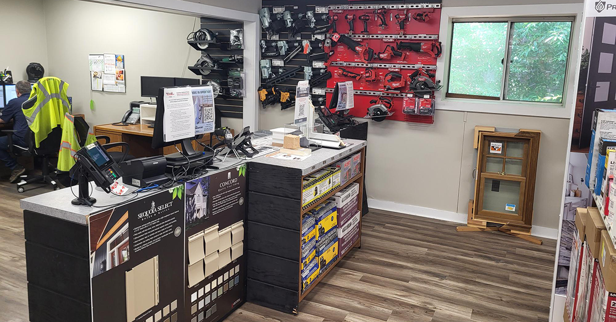 Interior showroom checkout counter with a point-of-sale terminal and computer monitors, flanked by wall-mounted nail guns and Milwaukee power tools. Siding color sample boards and a wood-framed window display sit alongside the counter, with a window overlooking greenery in the background.