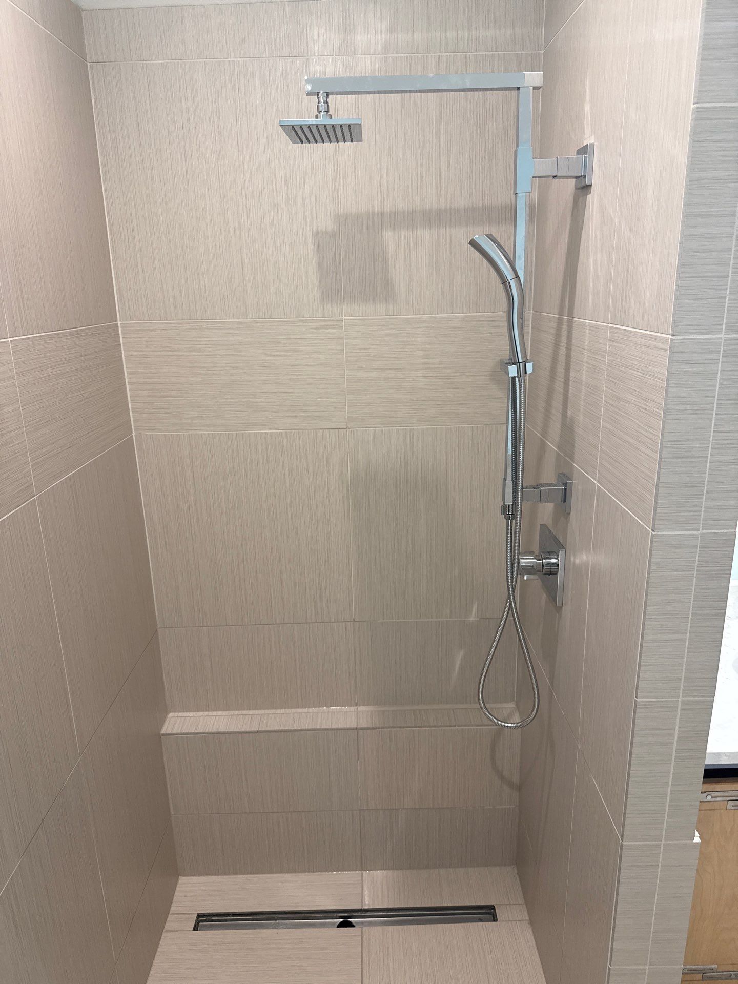 Tiled walk-in shower features a square overhead rainfall showerhead, handheld wand, chrome controls, beige wall and floor tiles, built-in bench, linear drain, and clean modern plumbing fixtures throughout the enclosure.
