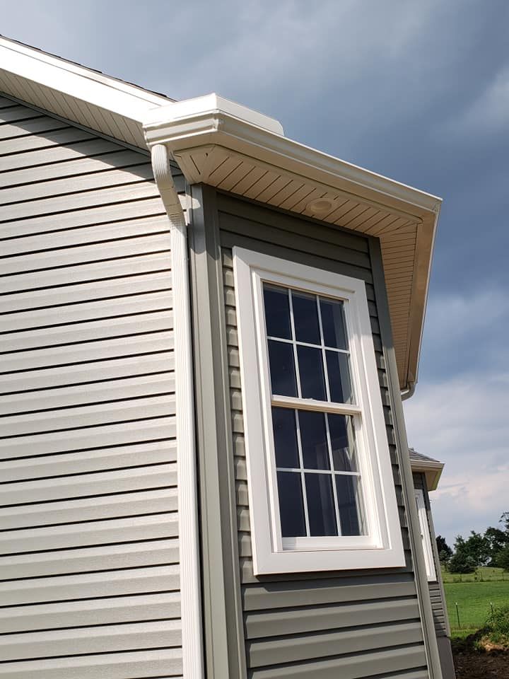 Close-up of a house exterior showing vinyl siding, a white-framed window, and a gutter system with downspout, highlighting clean installation and proper drainage design beneath the roofline.
