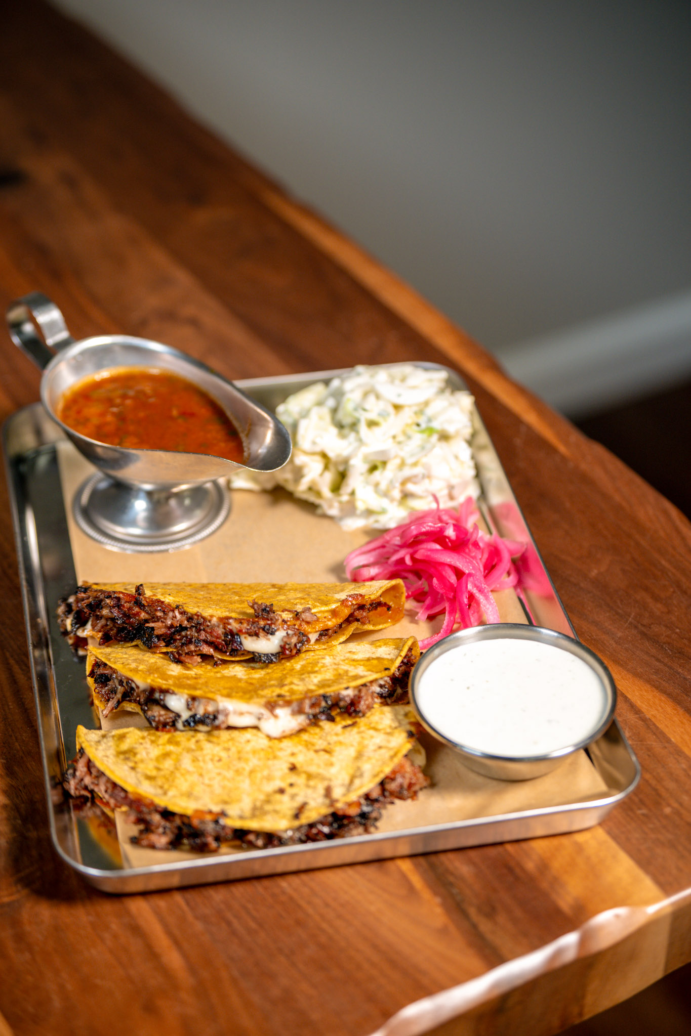 Three Smoked Beef Brisket Birria Tacos stacked on a metal tray with griddled corn tortillas, served with coleslaw, pickled red onions, serrano buttermilk ranch, and chipotle dipping broth.