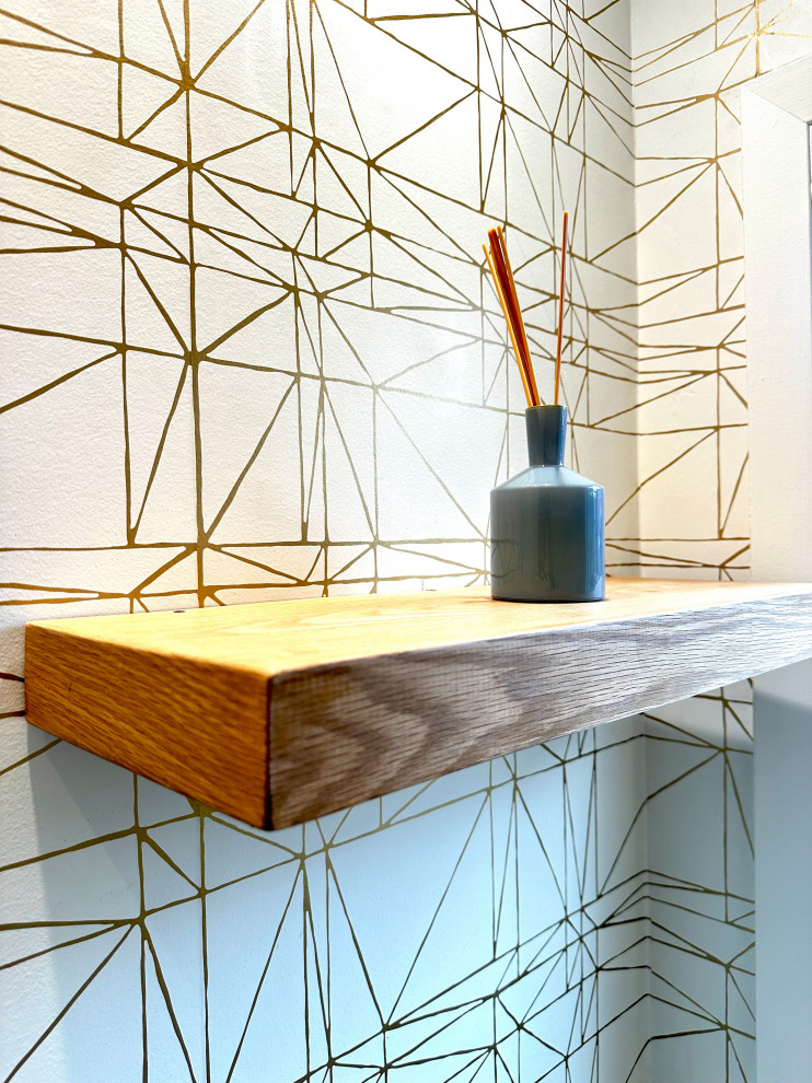 A close-up shot of a floating wooden shelf against a white wall with a geometric gold pattern. A small grey vase with reeds is placed on the shelf.