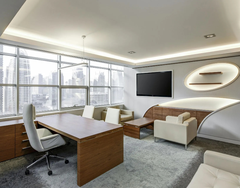 A modern corporate executive office featuring floor-to-ceiling windows with a city skyline view, a rich wood desk with a high-back desk chair, leather armchairs, a wall-mounted TV, and custom lit wall recessed shelves.
