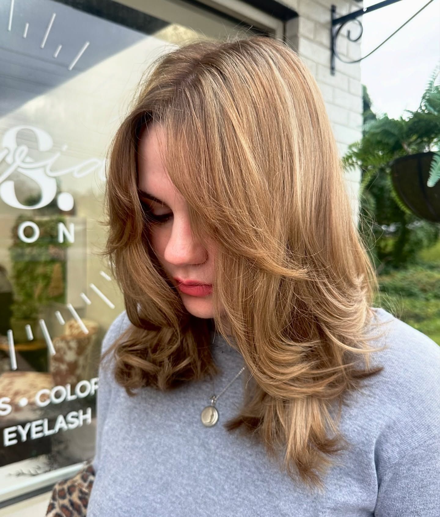 Woman with shoulder length layered light brown hair styled in soft blowout waves standing outside salon entrance.