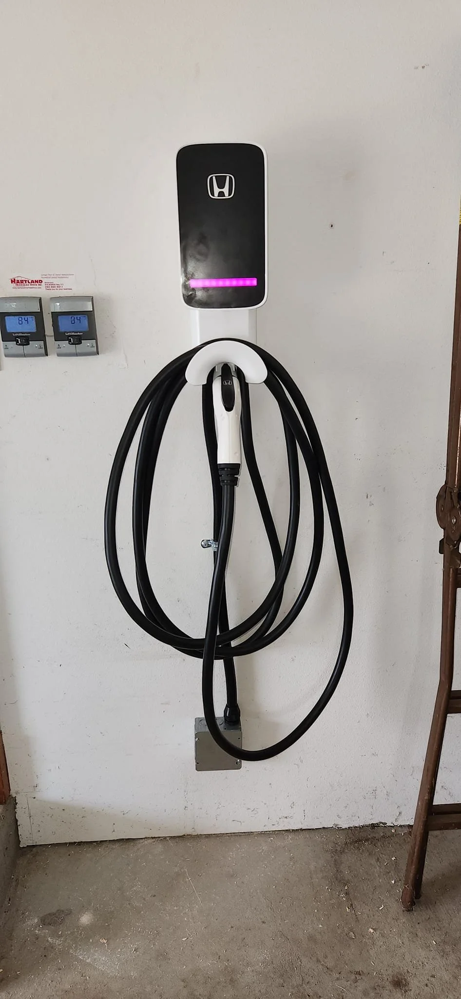 A Honda electric vehicle charger is mounted on a white wall, with a thick black charging cable coiled and hanging from a white hook. Below the charger, two gray digital displays show "84". A metal junction box is visible near the bottom of the cable. To the right, a portion of a wooden ladder is seen.