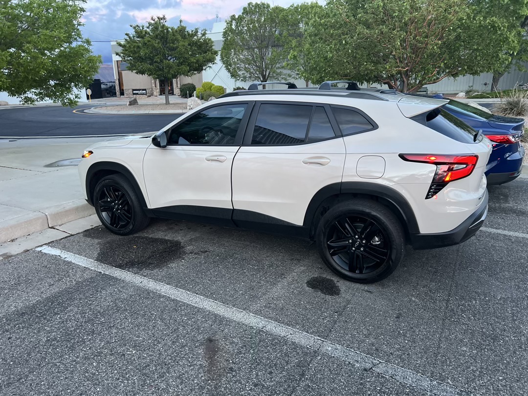 A white SUV with black rims parked in a parking lot with a blue SUV behind it. The SUV has tinted windows and a sleek design. The background shows a building with trees and a cloudy sky.