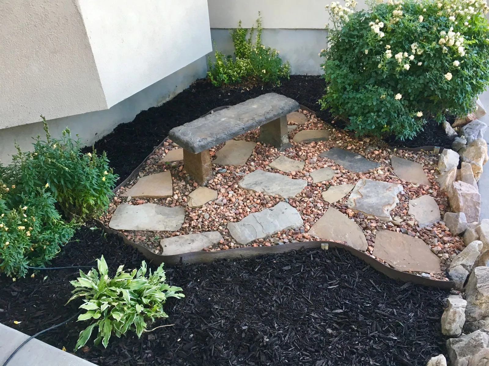 Decorative rock garden featuring natural stone bench, stepping stones, flowering shrubs, mulch, and  