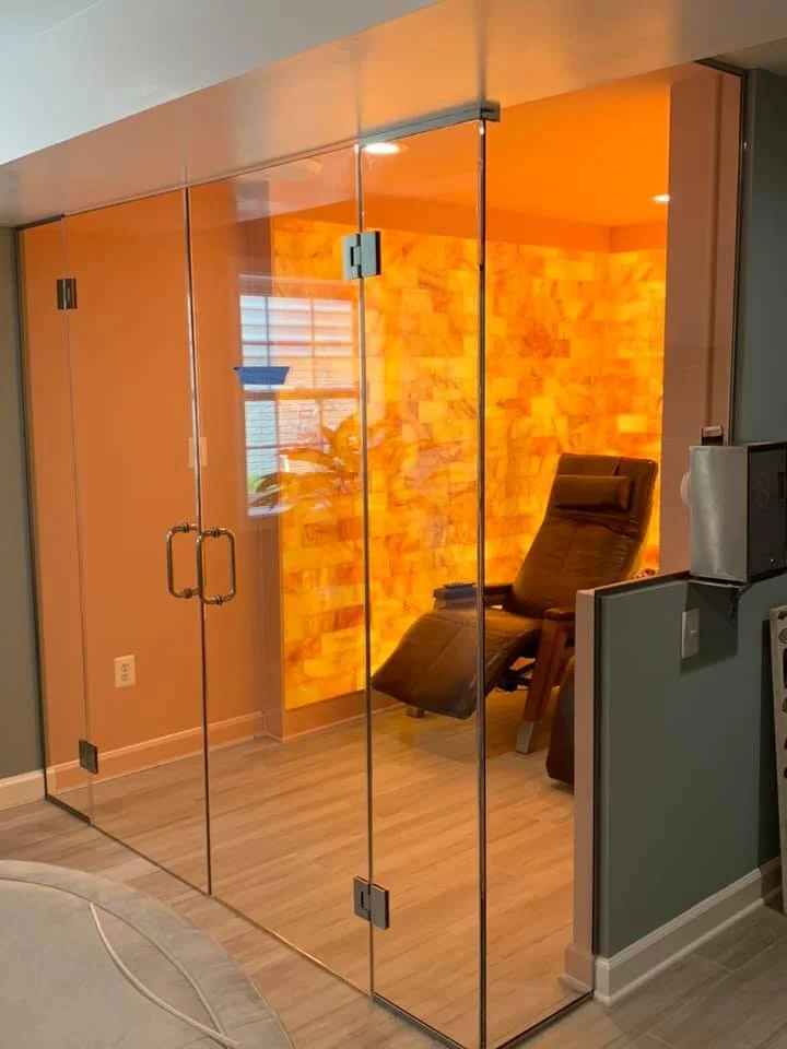 A salt cave room with a reclining chair and illuminated salt walls is visible through a glass enclosure. The room has wooden floors and an orange hue from the salt wall lighting. The enclosure has glass doors with chrome handles.