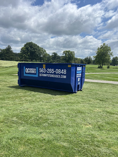 An empty blue QC Dumpster Dudes dumpster sits on a manicured green lawn, ideal for landscaping or outdoor cleanup projects.