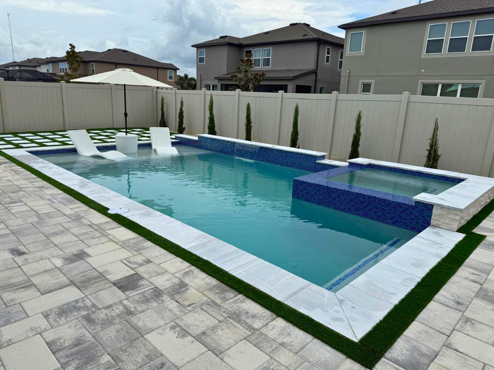 Modern backyard swimming pool with an attached raised spa, sun shelf with in-pool lounge chairs, artificial turf, and a white privacy fence.