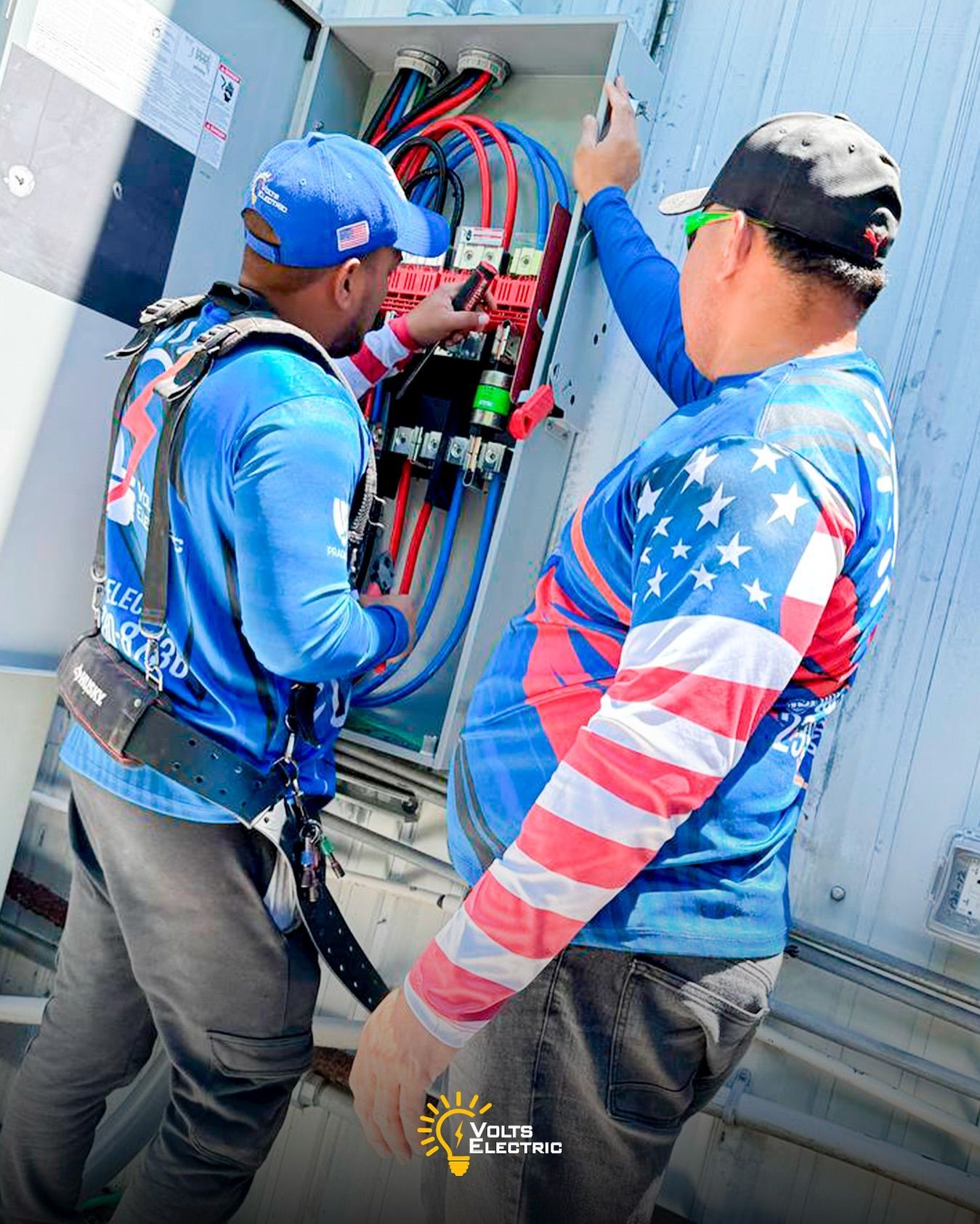 Two electricians inspecting and working inside an open outdoor electrical panel filled with heavy power cables and components, performing maintenance or installation while wearing branded Volts Electric uniforms.