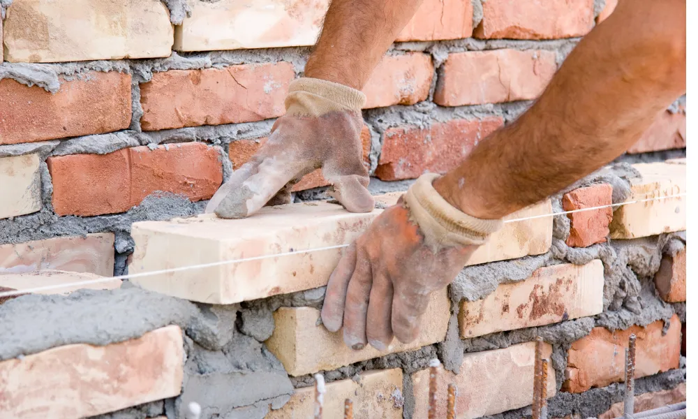 A close-up shot shows a bricklayer's hands, wearing work gloves, carefully placing a light-colored brick onto a wall under construction. Mortar is visible between the bricks, and a taut string line guides the placement. The wall is made of red and beige bricks, indicating ongoing construction work.