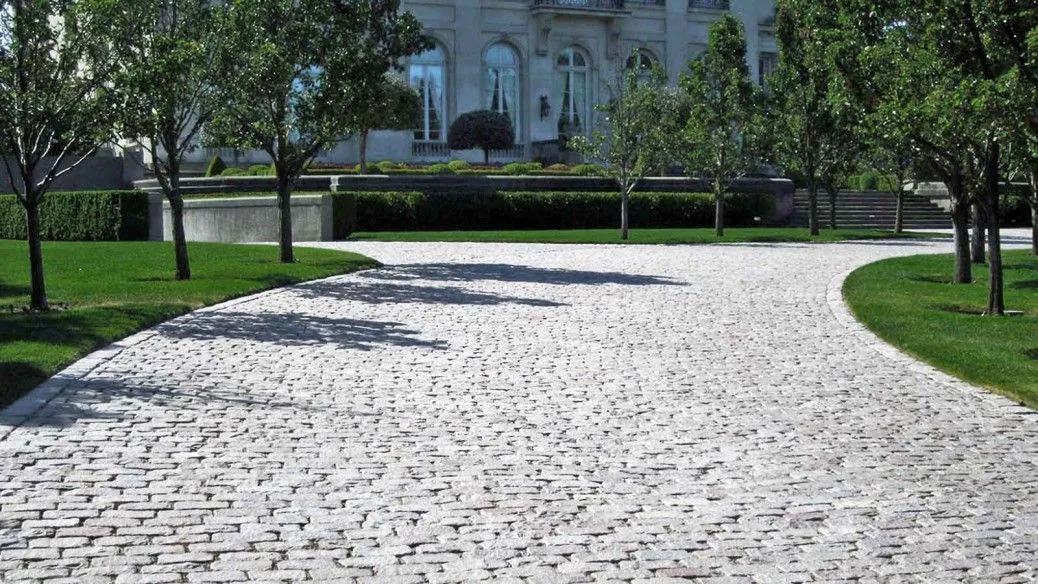 A curved cobblestone driveway leads to a grand building, flanked by manicured lawns and trees. The building appears to be a stately mansion with large windows and classical architecture.