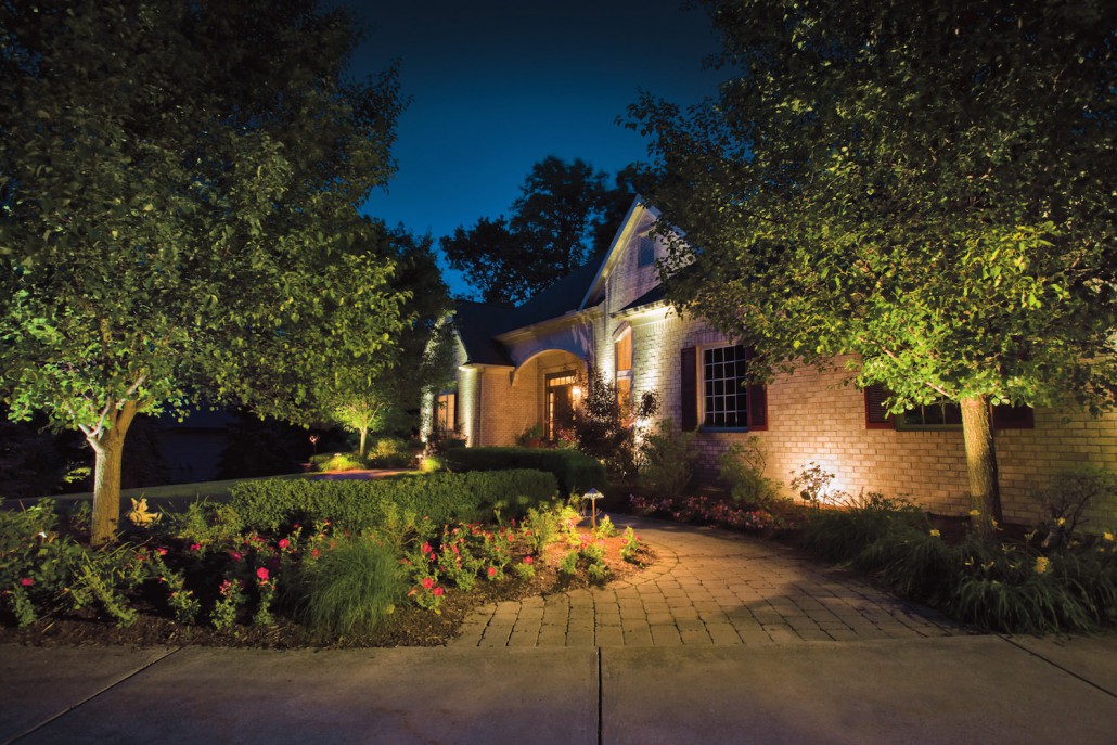 Nighttime view of an illuminated brick home, surrounded by lush trees and colorful flowerbeds, with a welcoming paved walkway and warm lights.
