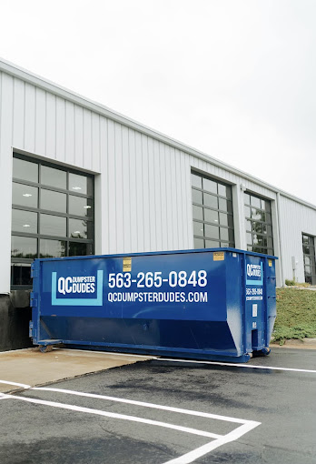 A large blue dumpster with QC Dumpster Dudes branding parked in front of a modern commercial building.