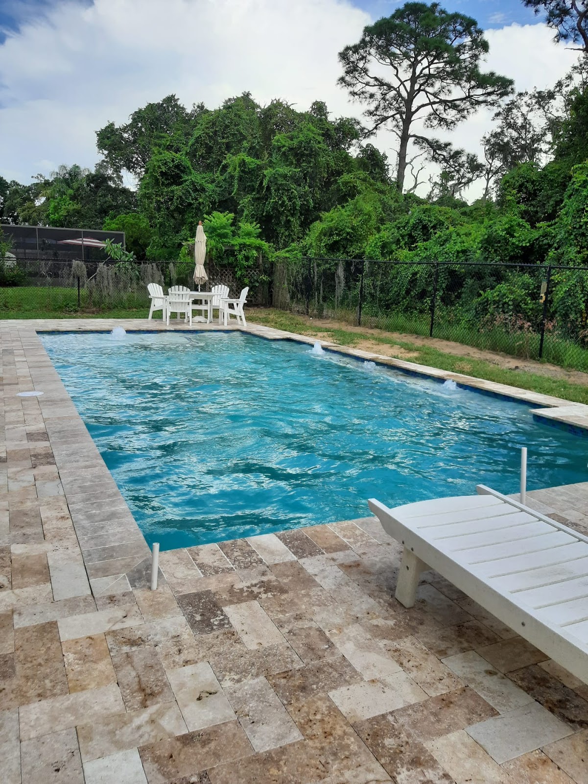 Rectangular residential swimming pool with clear blue water, light stone paver decking, lounge seating, and lush green landscaping.