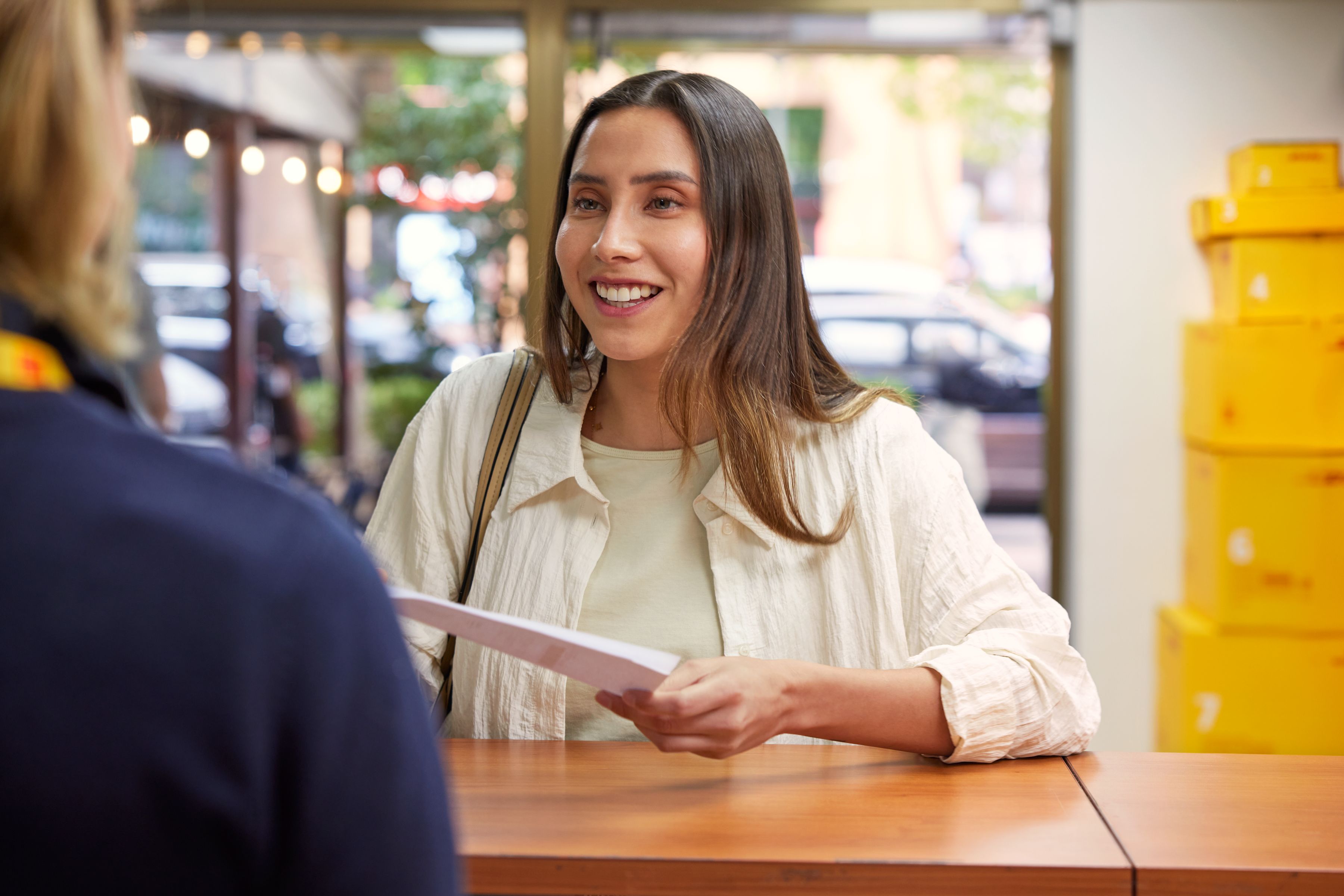 A customer is at a DHL Service Point counter holding a document while speaking with a staff member. Yellow DHL parcels are stacked in the background, and the setting appears to be a bright, modern retail location.