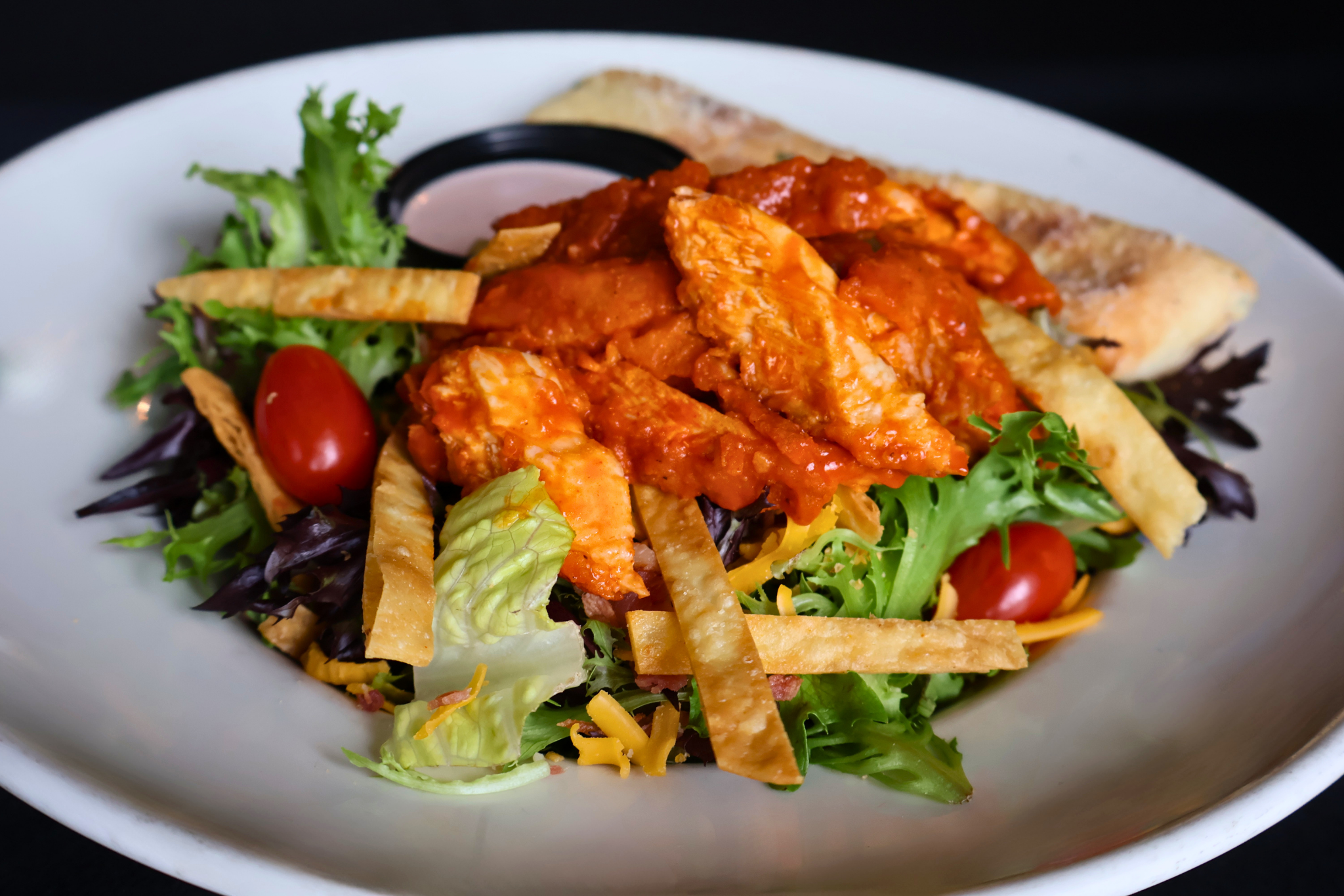 A fresh salad topped with saucy grilled chicken, crunchy tortilla strips, tomatoes, and cheese, served with a side of bread and dressing.