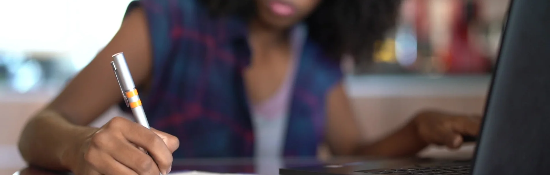 A young woman is shown from the chest down, writing with a pen in a notebook, while her other hand is on a laptop. She is wearing a dark blue and purple plaid vest over a light pink top. The background is out of focus with soft lighting.
