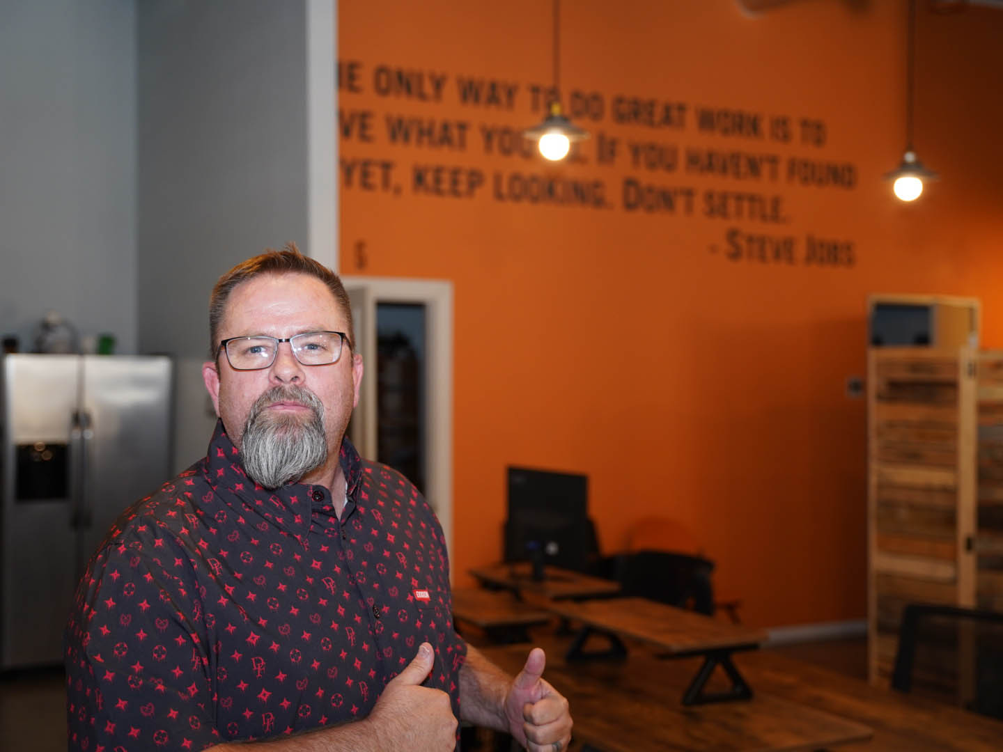 In this image, Mike Rux, a professional from Ciphers Digital Marketing, is seen indoors, giving two thumbs up with a confident and motivated expression. He is wearing glasses and a patterned short-sleeve shirt. The setting features an orange wall with a prominent motivational quote by Steve Jobs: "The only way to do great work is to love what you do. If you haven't found it yet, keep looking. Don't settle." The room is furnished with wooden tables and computers, creating a warm and inspiring workspace atmosphere at the Ciphers Digital Marketing office in Gilbert, Arizona.