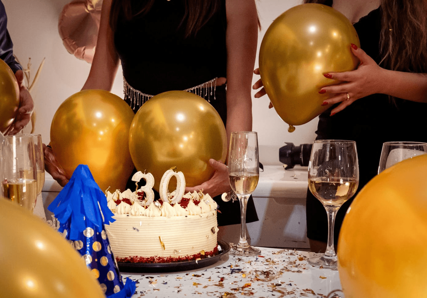 A close-up of a birthday cake with the number 30 on top, surrounded by gold balloons and champagne glasses on a table scattered with confetti. Three people, two women and a man, are partially visible holding balloons and dressed in party attire. The scene suggests a celebratory 30th birthday party.