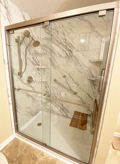 A spacious, modern shower stall with a marble-patterned wall and glass doors is equipped with a handheld showerhead, grab bars, and a wooden shower seat.