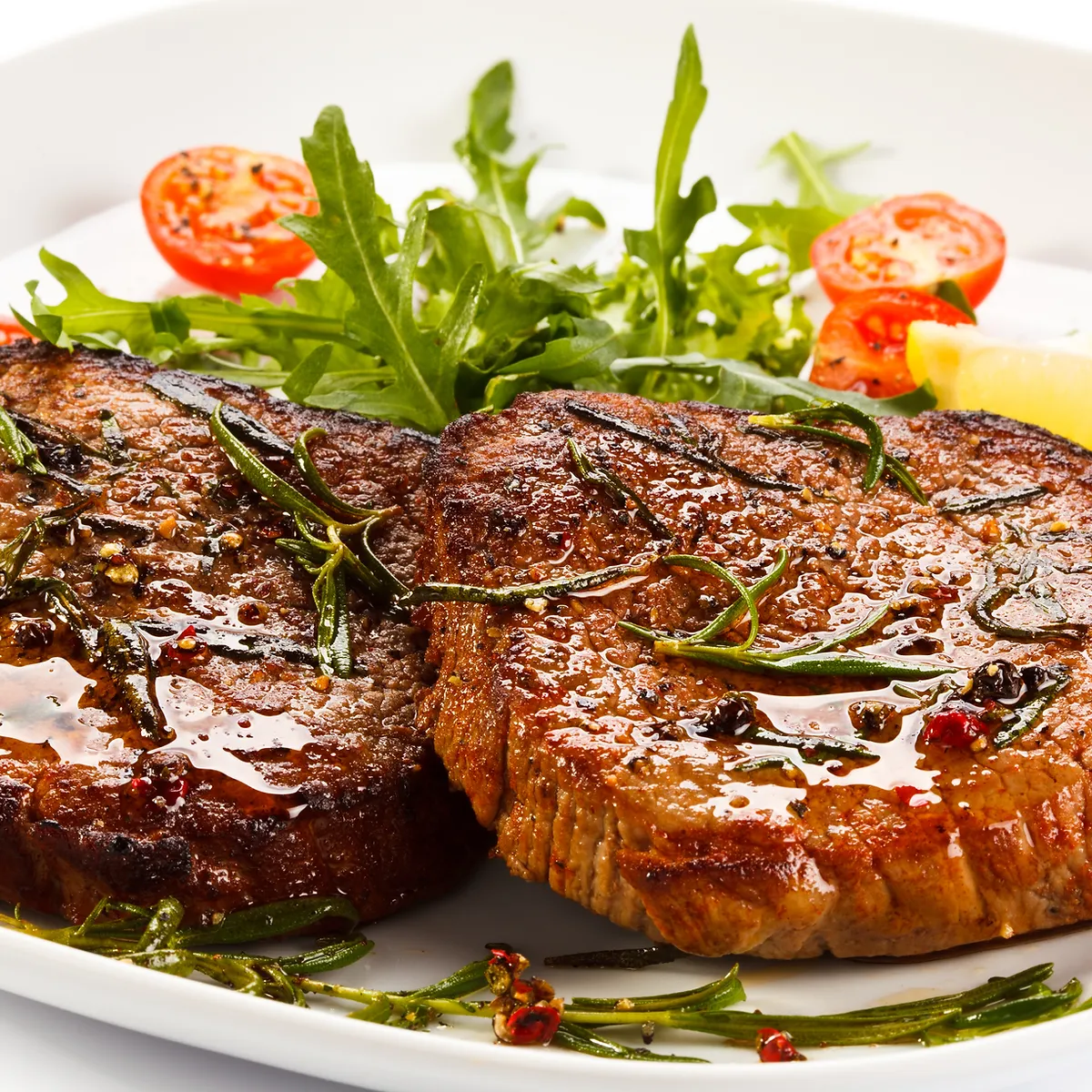 Two perfectly grilled steaks are served on a white plate, garnished with fresh rosemary sprigs, cherry tomatoes, and a wedge of lemon. The steaks are coated in a glistening sauce and seasoned with peppercorns and herbs, suggesting a rich and savory flavor profile. The vibrant green of the rosemary and arugula, contrasted with the deep brown of the steak and bright red of the tomatoes, creates an appealing visual presentation.