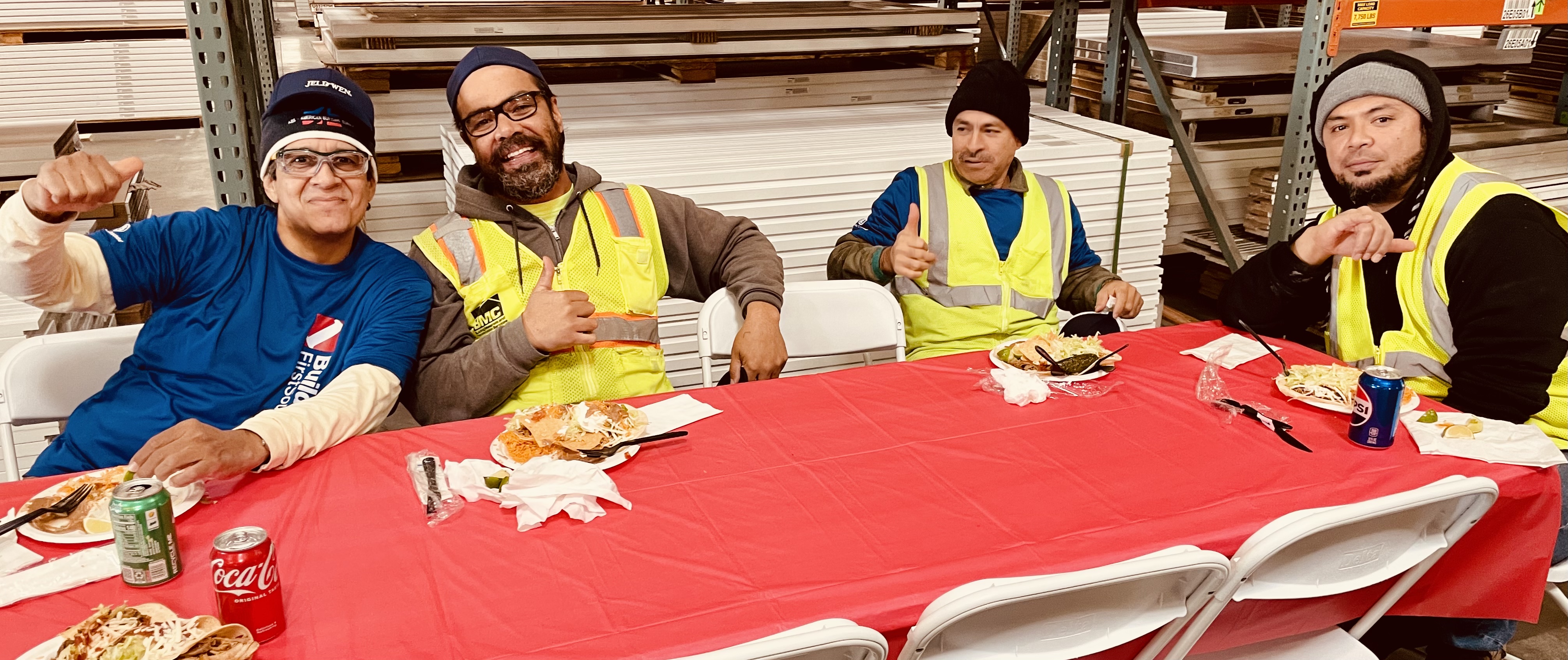 Four men sitting at a long table covered with a red tablecloth, enjoying a meal. They are smiling and showing thumbs-up gestures. They are wearing safety vests and casual clothing.