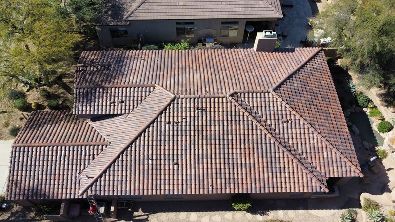 A top-down aerial drone view showcasing a complex residential roof featuring weathered brown and charcoal barrel tiles. The expansive home is set within a desert landscape with native greenery.