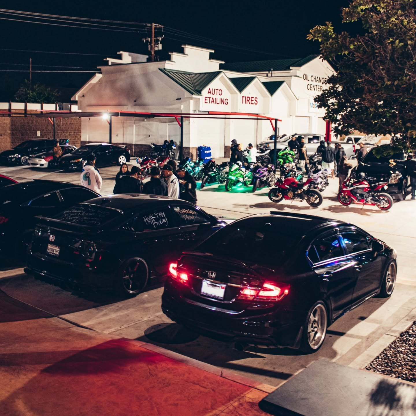 Large nighttime car and motorcycle meet taking place outside an automotive detailing and tire center 