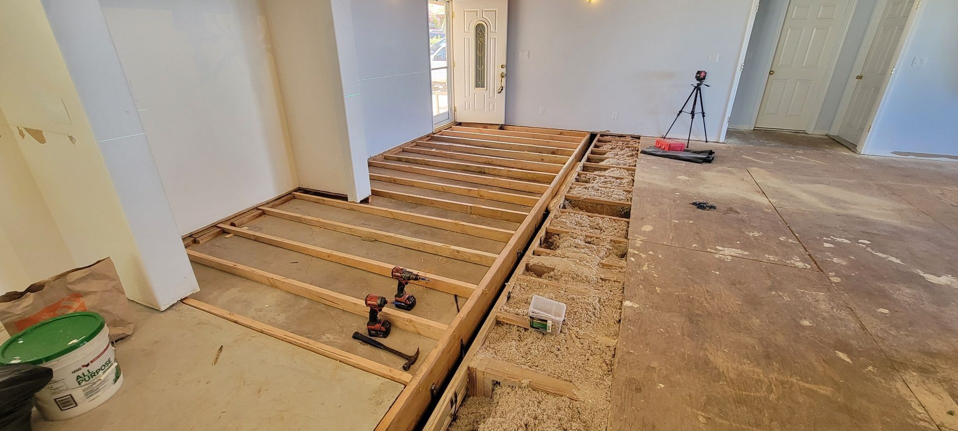 An interior floor is under construction, showing new wooden joists installed in sections to create different floor levels. Power drills and tools are resting on the framed sections near a front doorway.