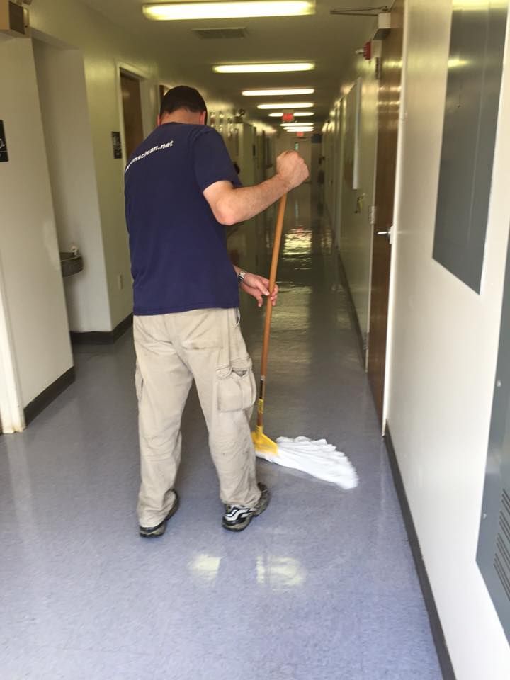 Janitorial worker mopping a long hallway floor in a commercial building, maintaining cleanliness and hygiene in a high-traffic indoor corridor with doors and lighting along both sides.