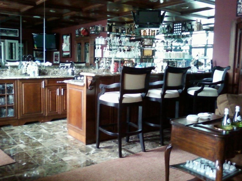 A stylish home bar area featuring wooden cabinetry, granite countertops, and high bar stools. Glass shelves display bottles and glassware, with mounted TVs and warm lighting creating a cozy entertainment space.