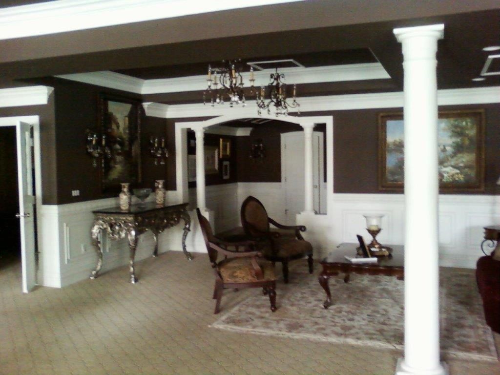 An elegant living area featuring decorative columns, chandeliers, and classic furniture. Dark walls contrast with white trim, while artwork and ornate table accents create a refined and luxurious interior atmosphere.