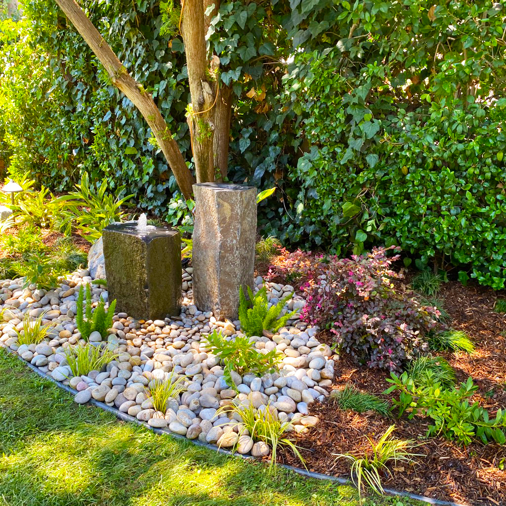 Lush garden with two stone fountains, smooth river rocks, ornamental grasses, purple shrubs, and dense green foliage in bright sunlight.