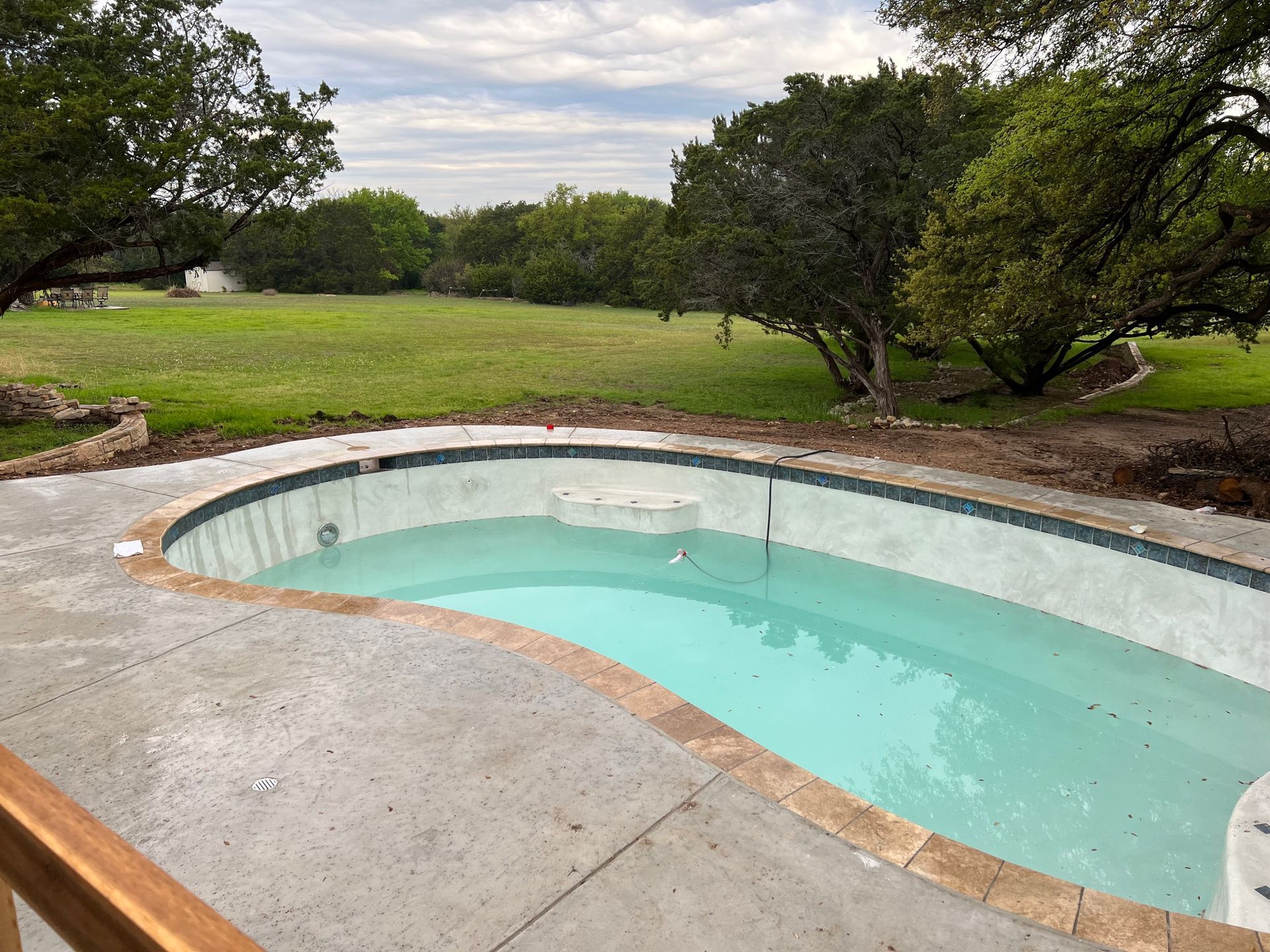 A nearly completed inground pool is being filled with fresh water. The design features tan tile coping, a decorative tile waterline, and a smooth concrete deck overlooking a grassy landscape.