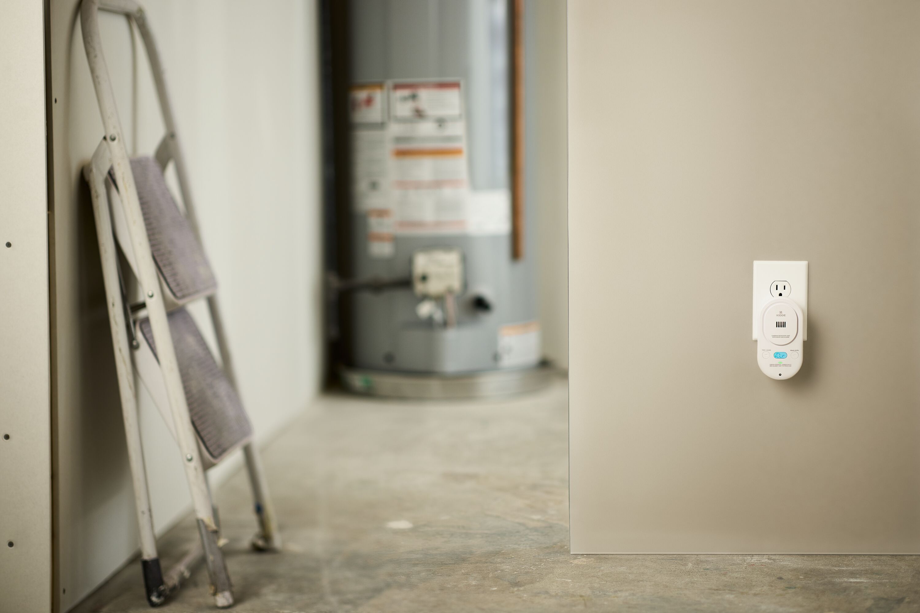 An image of a CO detector plugged into the wall, a step-stool and a water heater is in the background.