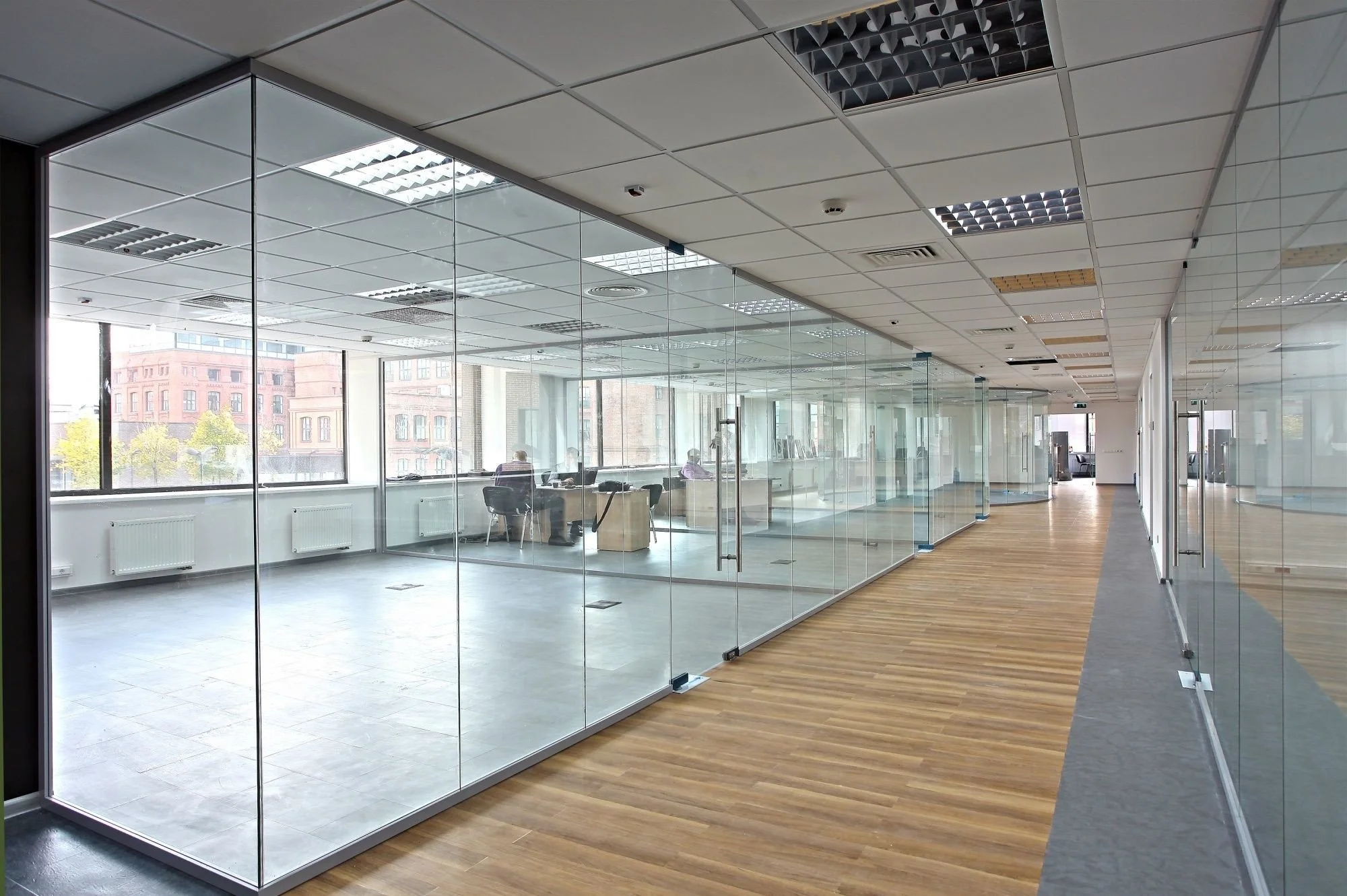 A modern office interior with glass partitions, wooden floors, and an illuminated ceiling. Several workstations are visible, some occupied by people working at computers, and a long hallway stretches into the distance.