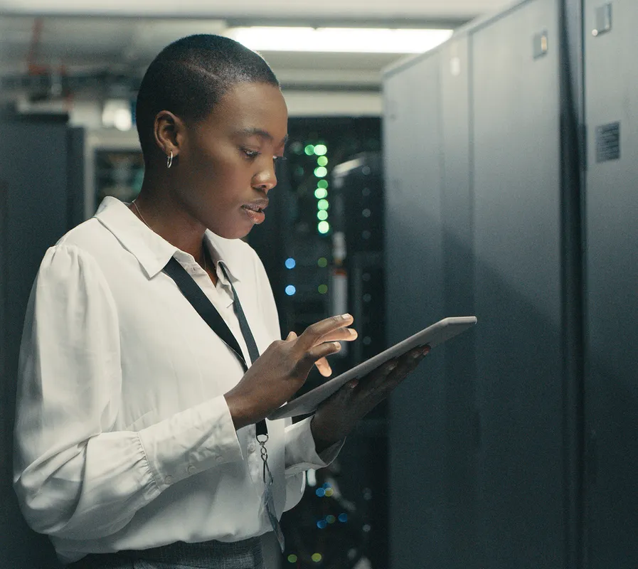 A Black woman with a buzzed haircut and hoop earrings, wearing a white button-down shirt and a lanyard, stands in a server room, looking down and interacting with a tablet. She is surrounded by tall, dark server racks, some of which have illuminated green and blue lights, suggesting active machinery.