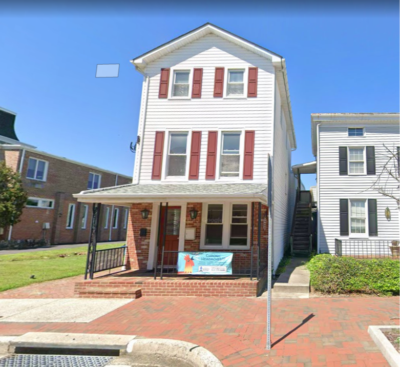 It&rsquo;s a bright daytime street-view style photo of a tall, narrow two-story building with white siding and red shutters. The lower level has a brick front with a small covered porch and black railings leading up a few steps.
There&rsquo;s a red front door and windows along the porch, plus a banner/sign displayed across the porch railing. To the right side of the building, there&rsquo;s an exterior staircase leading up to the second floor. The building sits along a brick sidewalk/entry path with neighboring buildings close on either side under a clear blue sky.