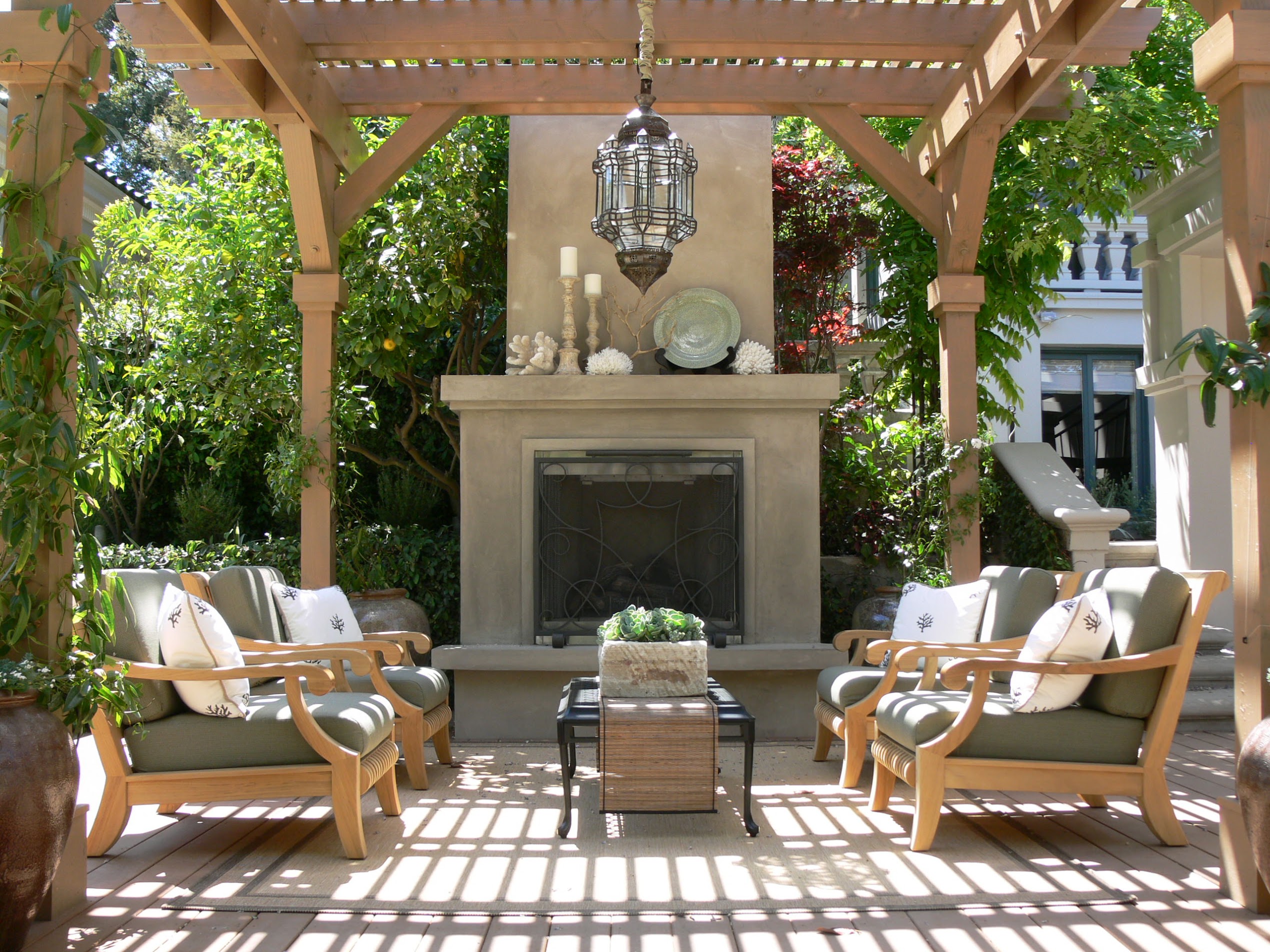 Sunlit outdoor patio with cushioned lounge chairs, stone fireplace, hanging lantern, lush greenery, and a warm, serene atmosphere.