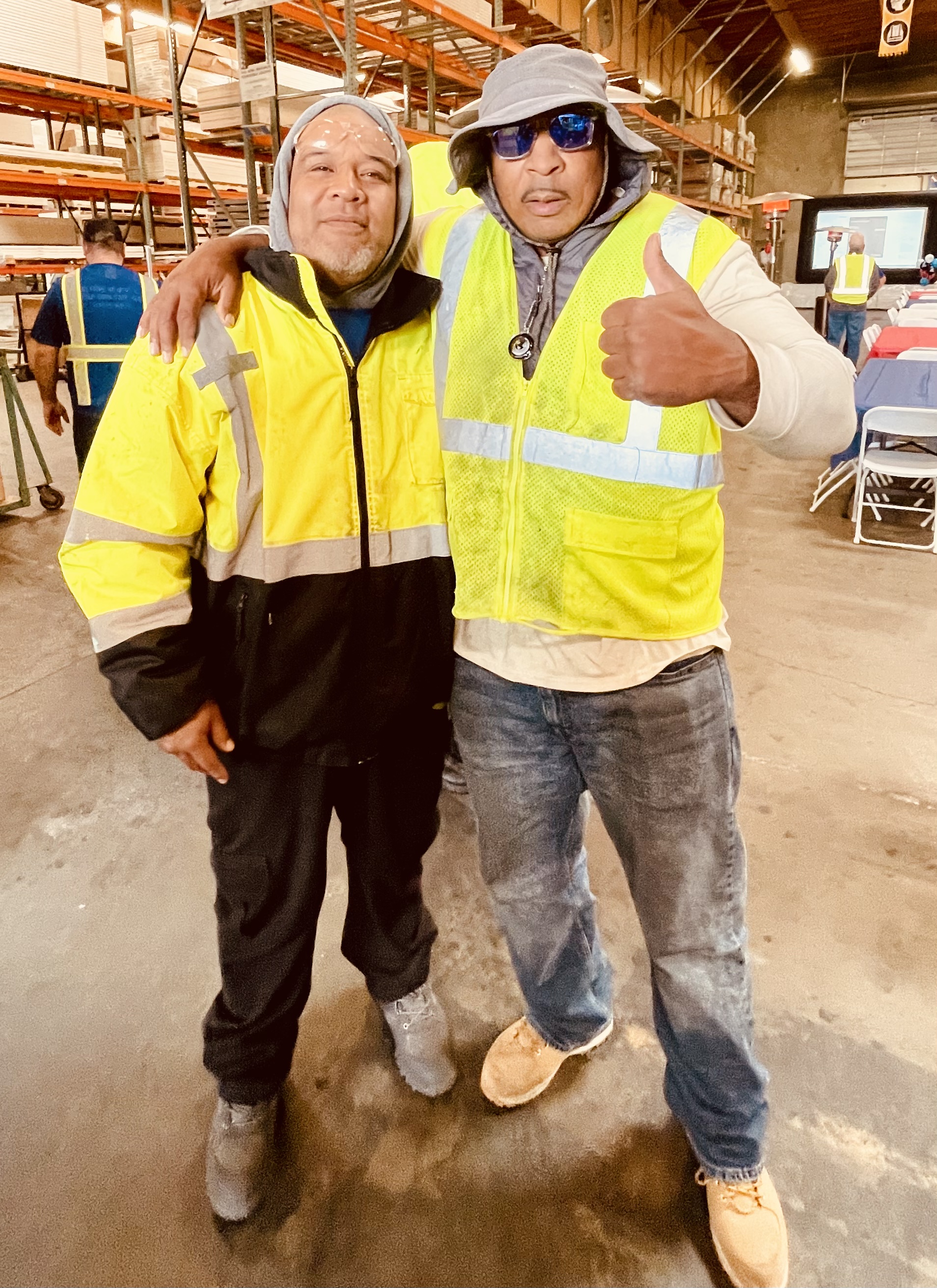 Two men wearing yellow safety vests and jackets are standing together in a warehouse, smiling and posing for the camera. The man on the right is giving a thumbs-up gesture.