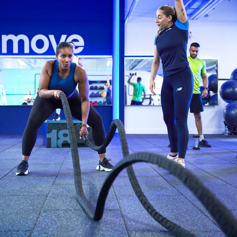 A woman in a blue tank top and black leggings is intensely engaged in a battle rope workout at the gym, while a trainer in navy blue offers guidance. The gym is brightly lit with mirrors, workout equipment, and other individuals exercising in the background.