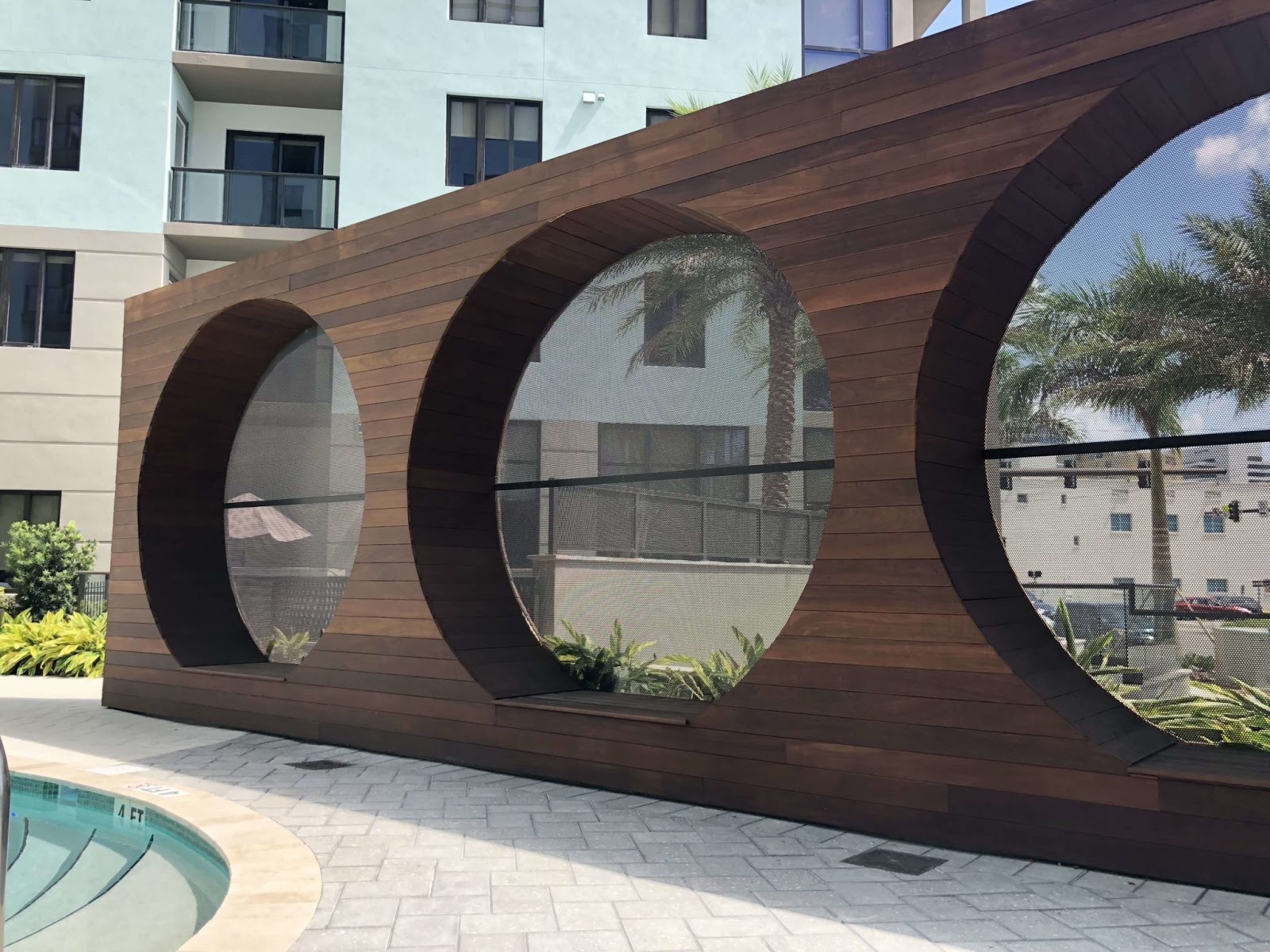Modern poolside architectural feature with curved wood-look panels and circular openings beside the swimming pool.