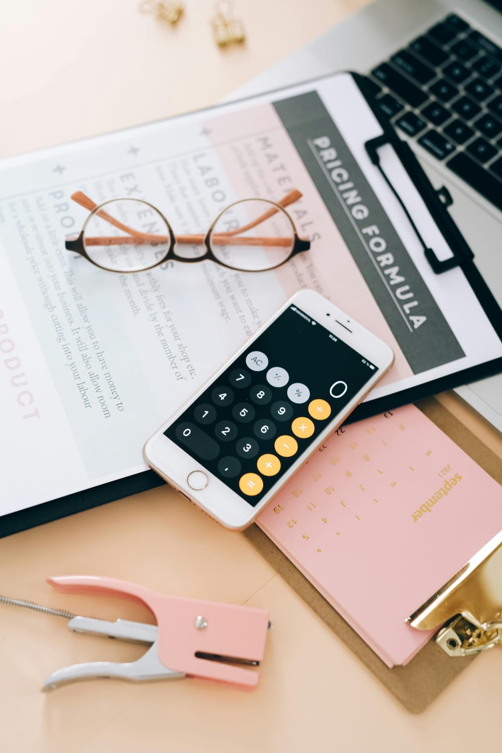 A flat lay of business and office supplies is arranged on a desk. The items include a pair of round, wire-rimmed glasses with light brown arms, a laptop, a clipboard with a "PRICING FORMULA" document, a smartphone displaying a calculator app, a pink stapler, and a pink calendar showing the month of September 2021. The background is a light peach color.