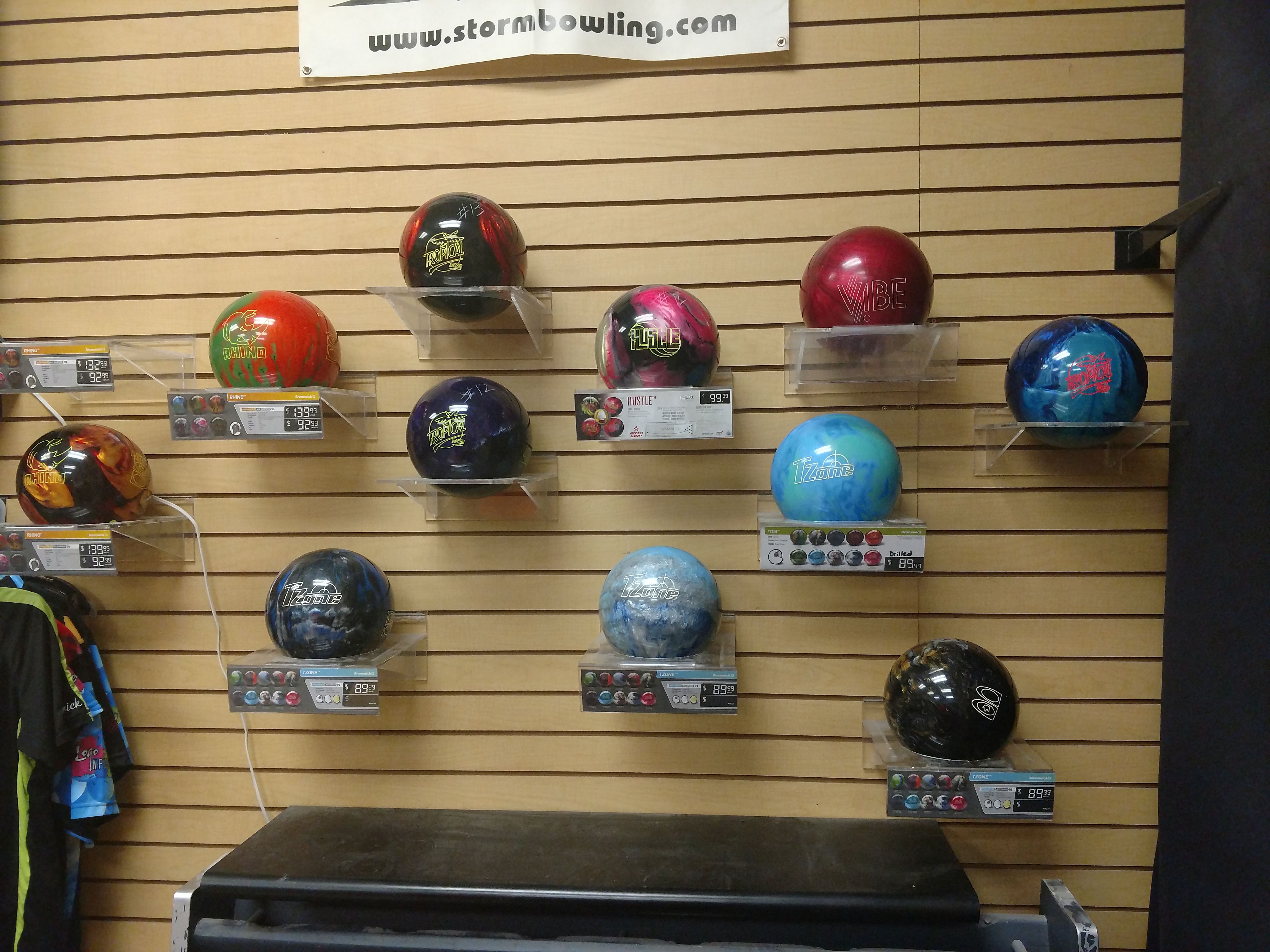 A display of bowling balls on a slatted wooden wall. The balls are of various colors and designs, and some have price tags and branding visible. A banner with a website address is displayed above the bowling balls.