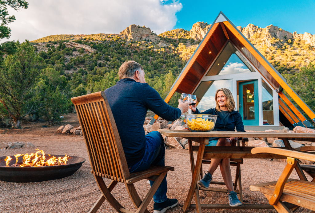 A couple enjoying a glass of wine and snacks outside of their triangular cabin near the mountains.