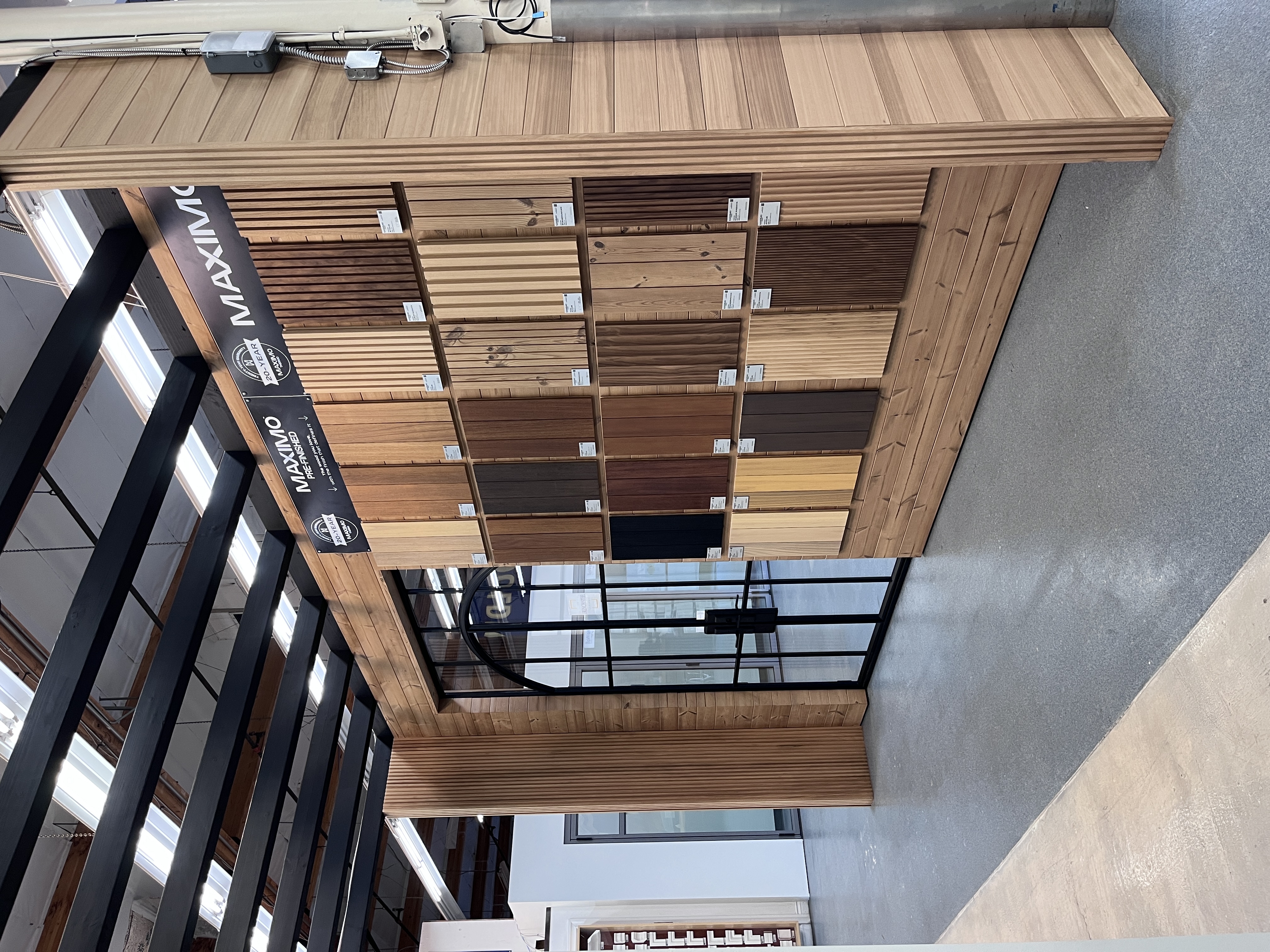 Maximo pre-finished decking and siding sample wall at Builders FirstSource Thousand Oaks, CA, showing a range of wood tones next to a steel-framed showroom door.