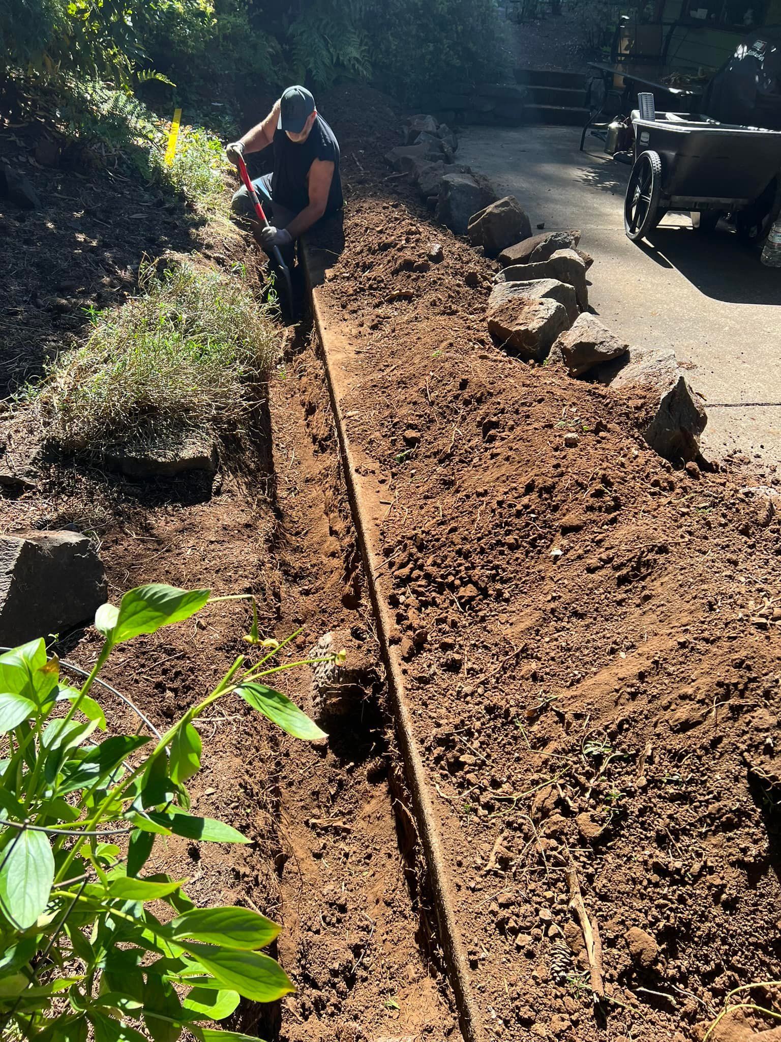 A worker in a black cap and sleeveless shirt digs a narrow trench with a red-handled shovel along a garden slope beside a driveway lined with large boulders.