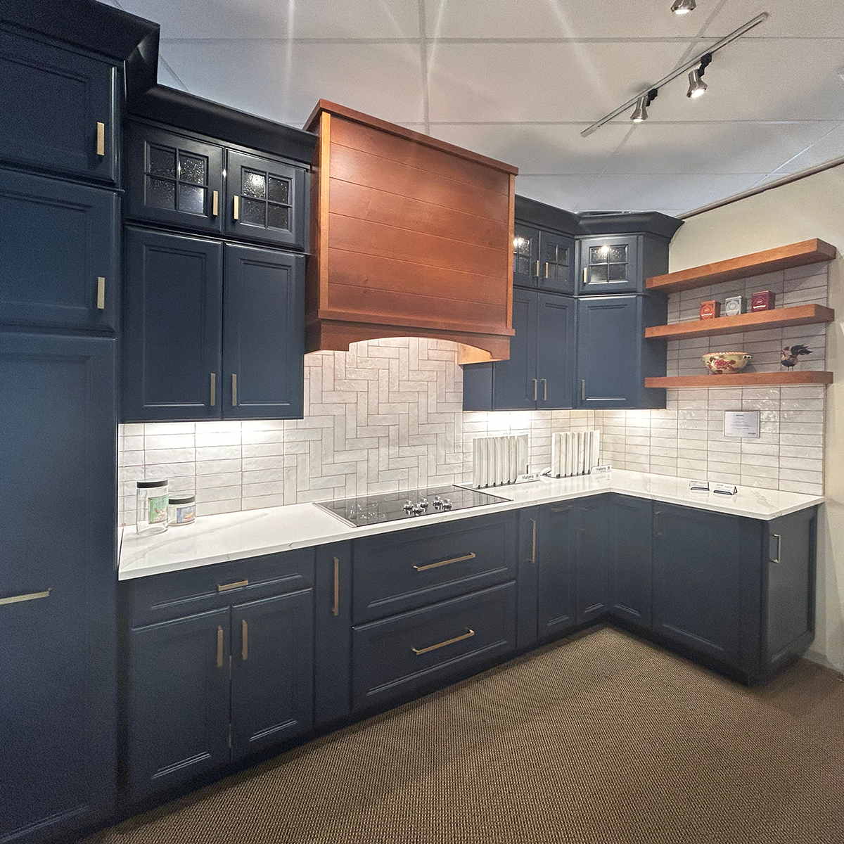 Kitchen showroom vignette featuring dark blue cabinetry with brass hardware, a white countertop and herringbone tile backsplash, wooden range hood, and floating wood shelves on the right wall.